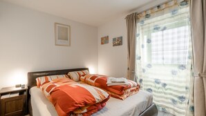 Una cama bien tendida con ropa de cama a rayas, una mesita de noche de madera con una lámpara y una ventana con cortinas florales.