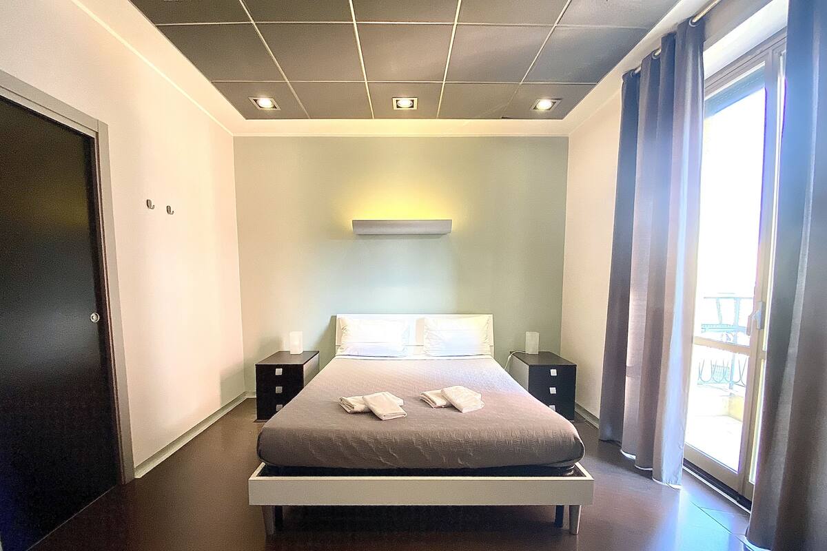 A modern hotel room with a large bed, bedside tables, a wardrobe, and a window with curtains.