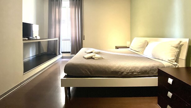 A hotel room with a bed, a TV on a stand, a nightstand, and a window with curtains. - Gemini Guest House (Rome)
