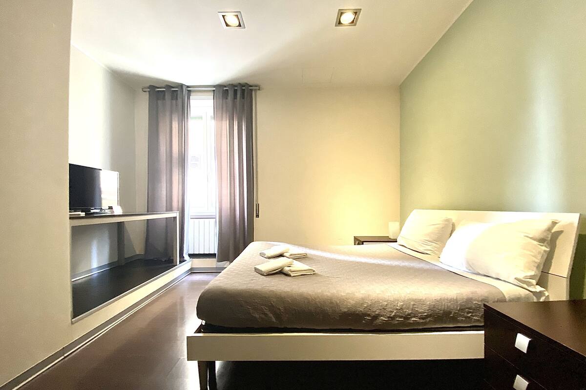 A hotel room with a bed, a TV on a stand, a nightstand, and a window with curtains.