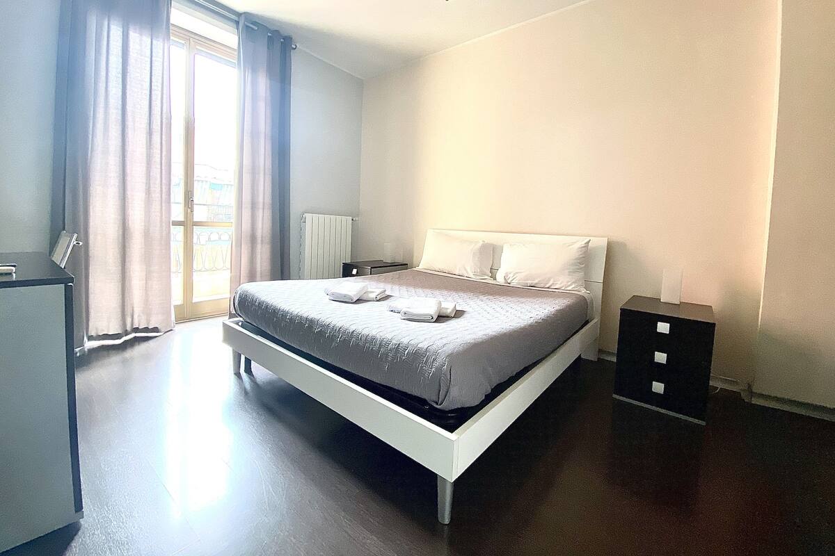 A bedroom with a bed, a desk, a radiator, and a window with curtains.