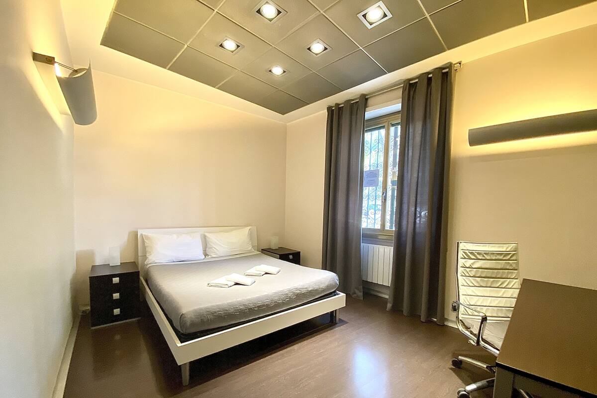 A modern bedroom with a bed, bedside tables, a desk, and a window with curtains.