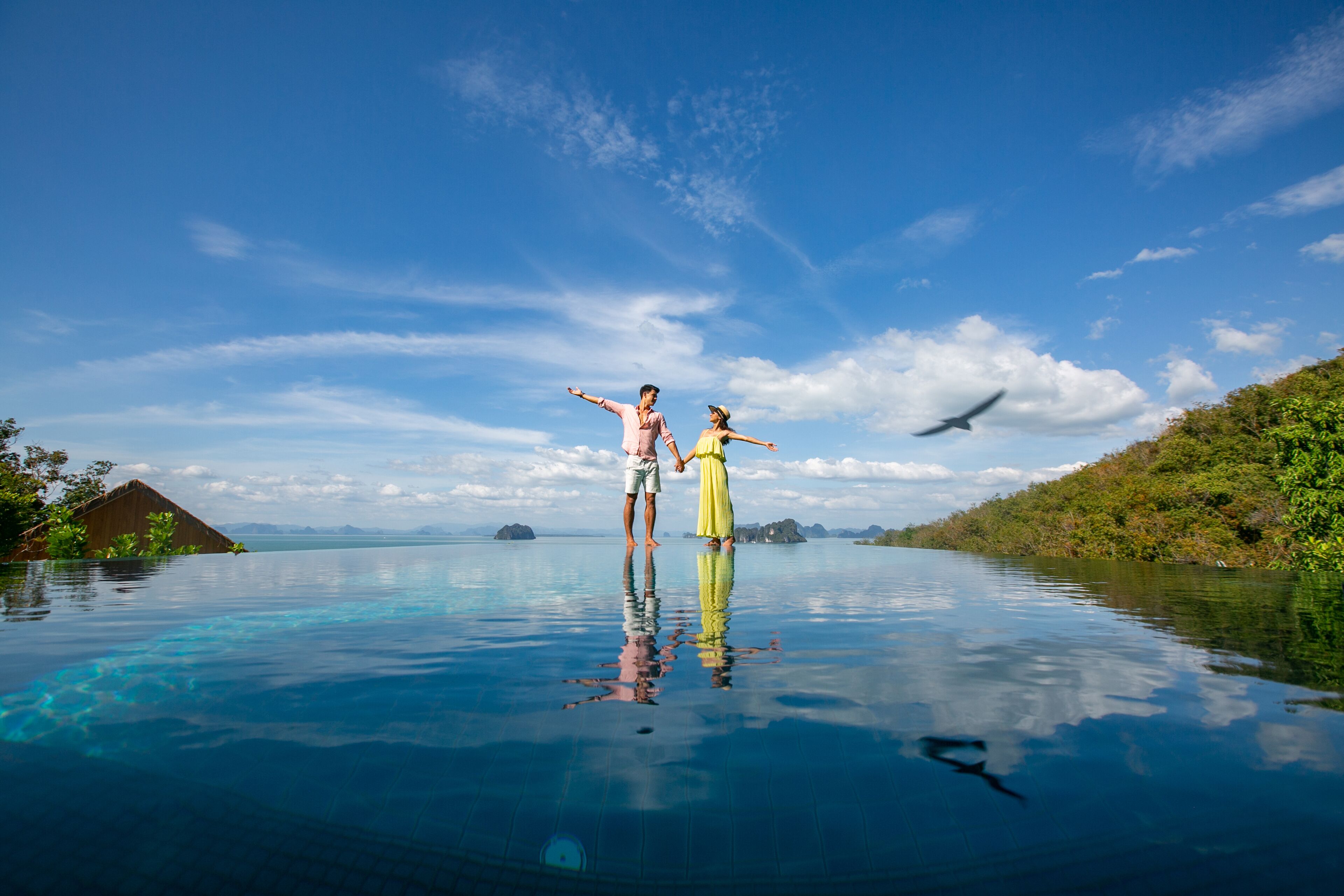 Una pareja parada en una piscina infinita azul cristalina, con vista al océano y a islas distantes.