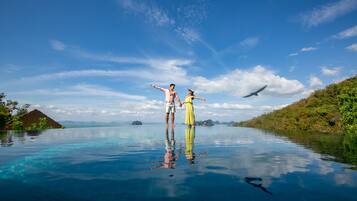 Una pareja parada en una piscina infinita azul cristalina, con vista al océano y a islas distantes.