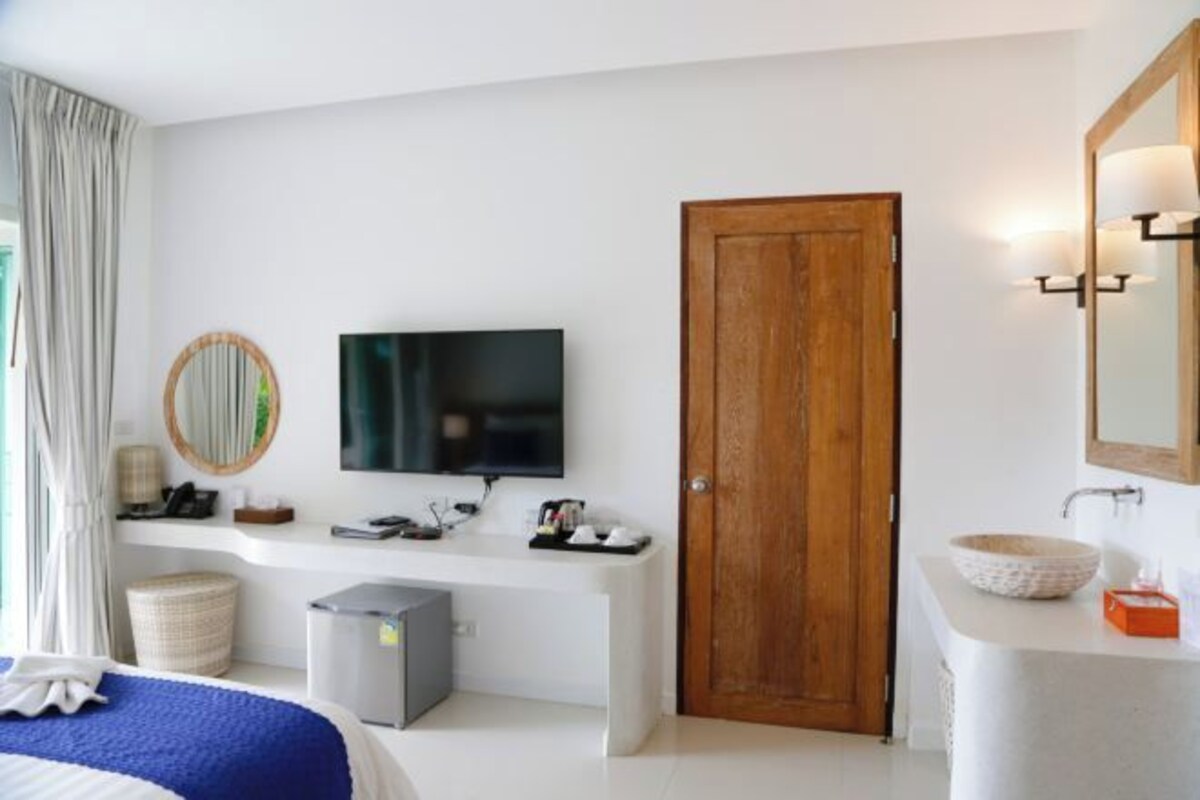 A hotel room with a bed, a TV, a wooden door, a white console table, a bathroom with a sink and mirror, and a window with curtains.