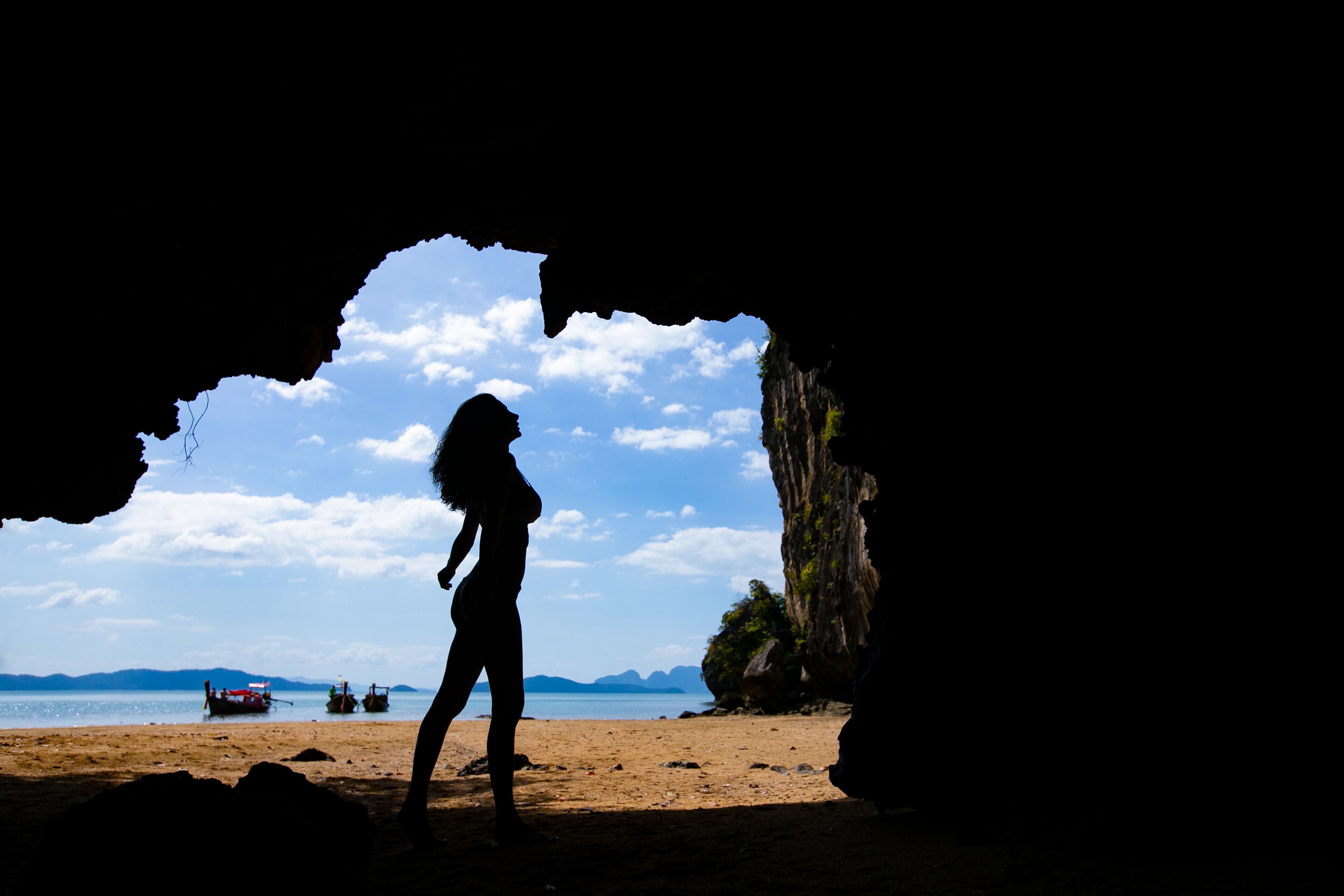 A silhouette of a person standing in a cave opening with a beach and boats in the background.