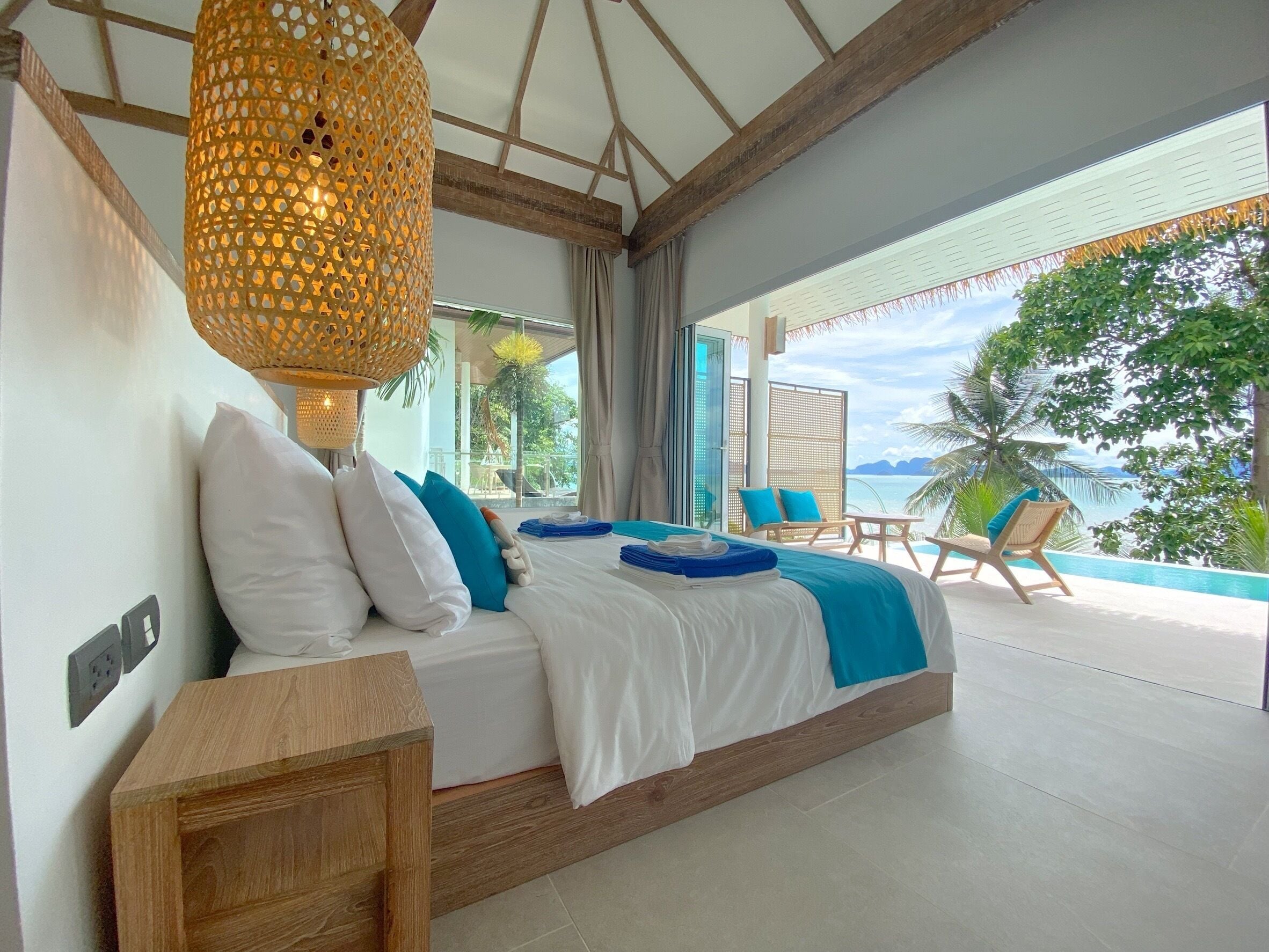 A bedroom with a large bed, a wooden nightstand, a hanging light fixture, and a view of the ocean.