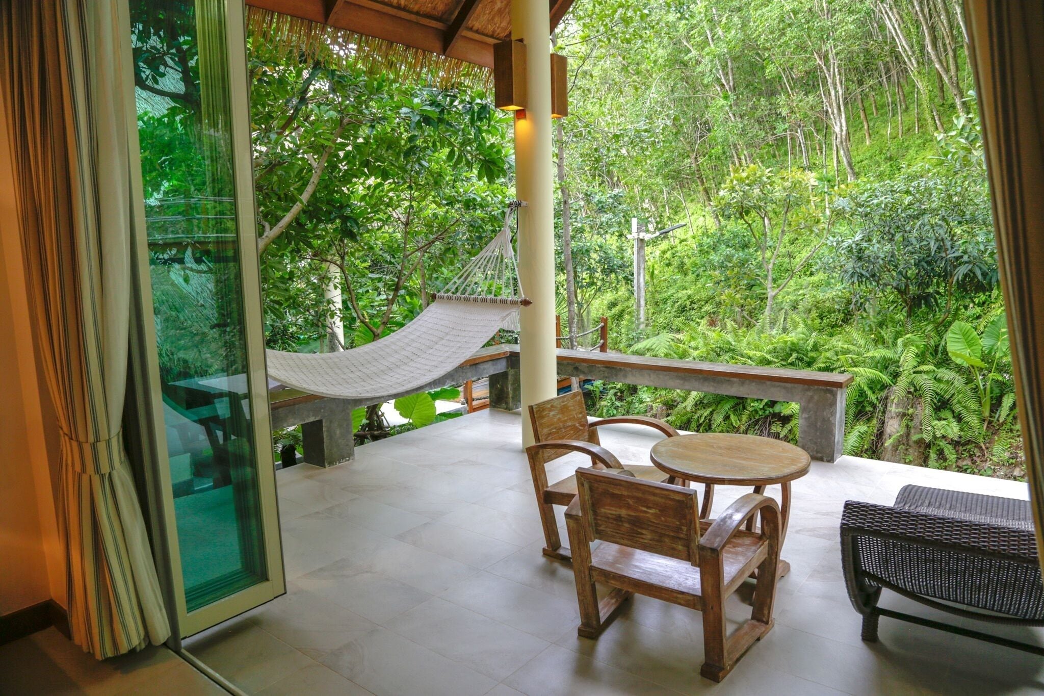 A balcony with a hammock, wooden chairs, and a table, overlooking a lush green forest.