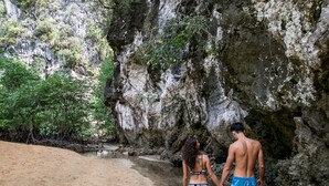 Una pareja camina sobre una playa arenosa, con un acantilado rocoso al fondo.