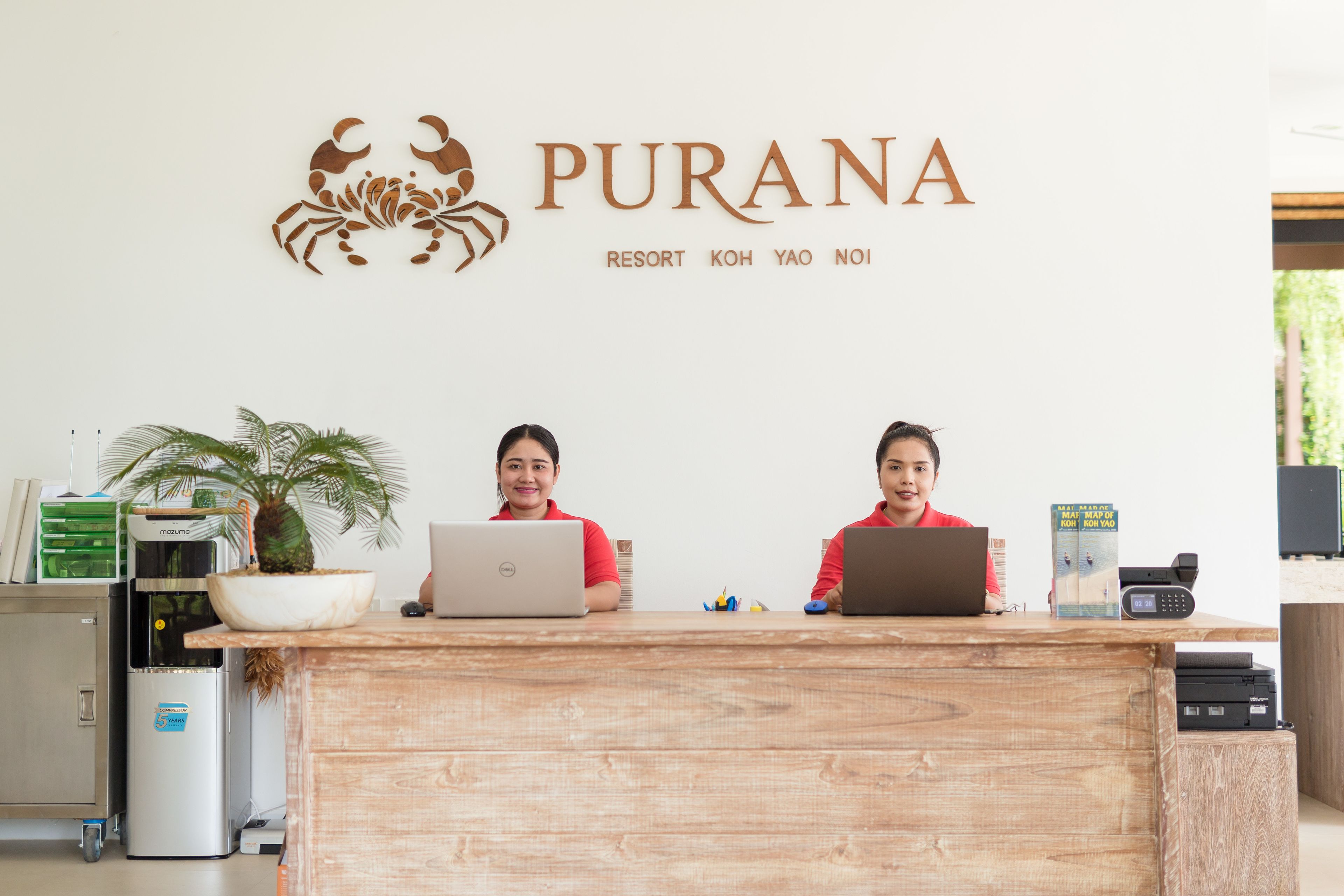 A reception desk with two staff members, a logo above them, and a plant to the left.