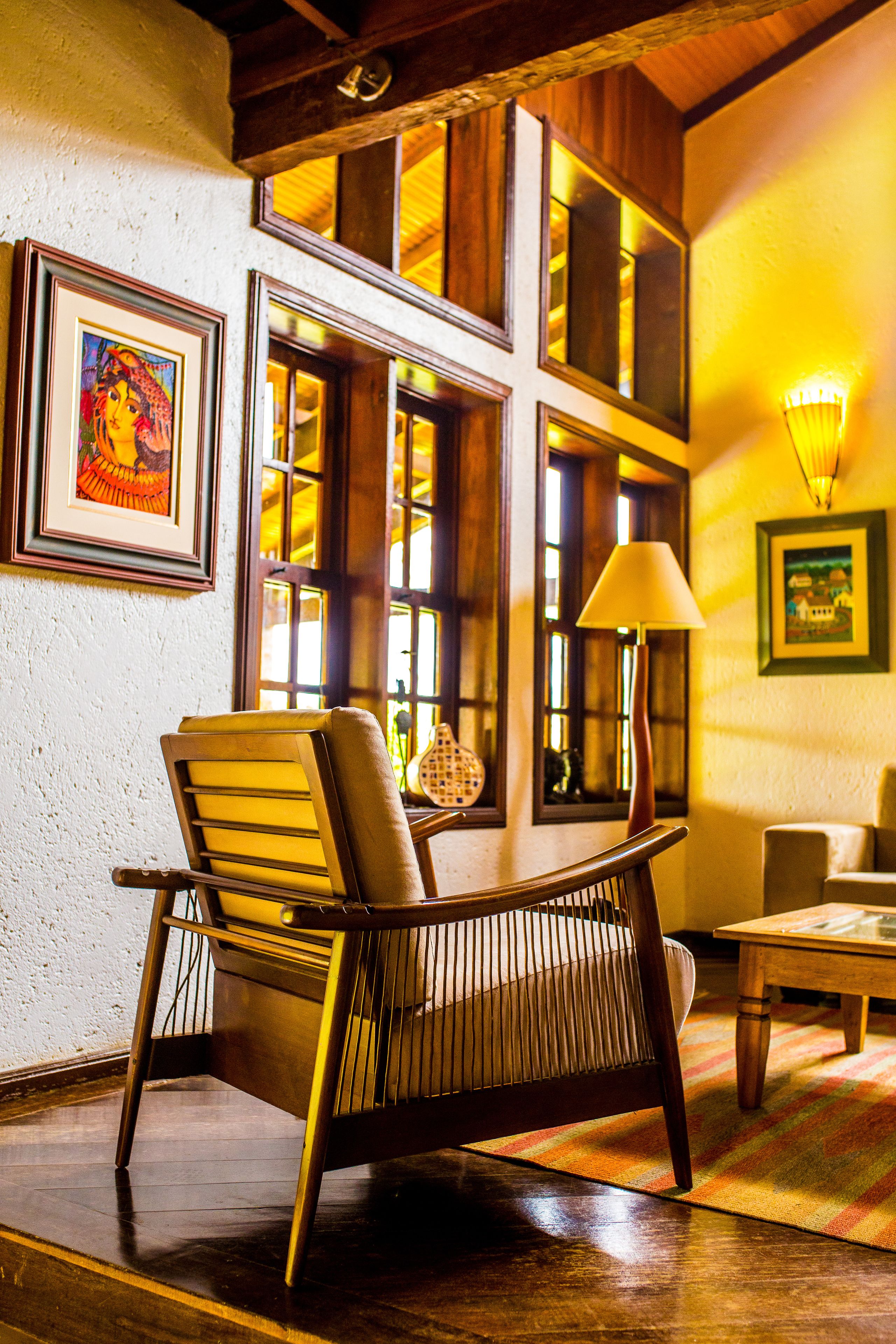 A room with a wooden chair, a table, and a lamp. There are windows with curtains and framed artwork on the walls.