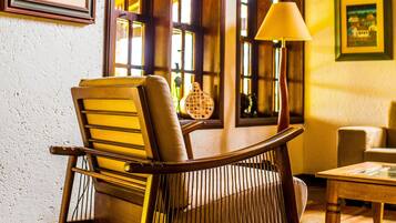 A room with a wooden chair, a table, and a lamp. There are windows with curtains and framed artwork on the walls.