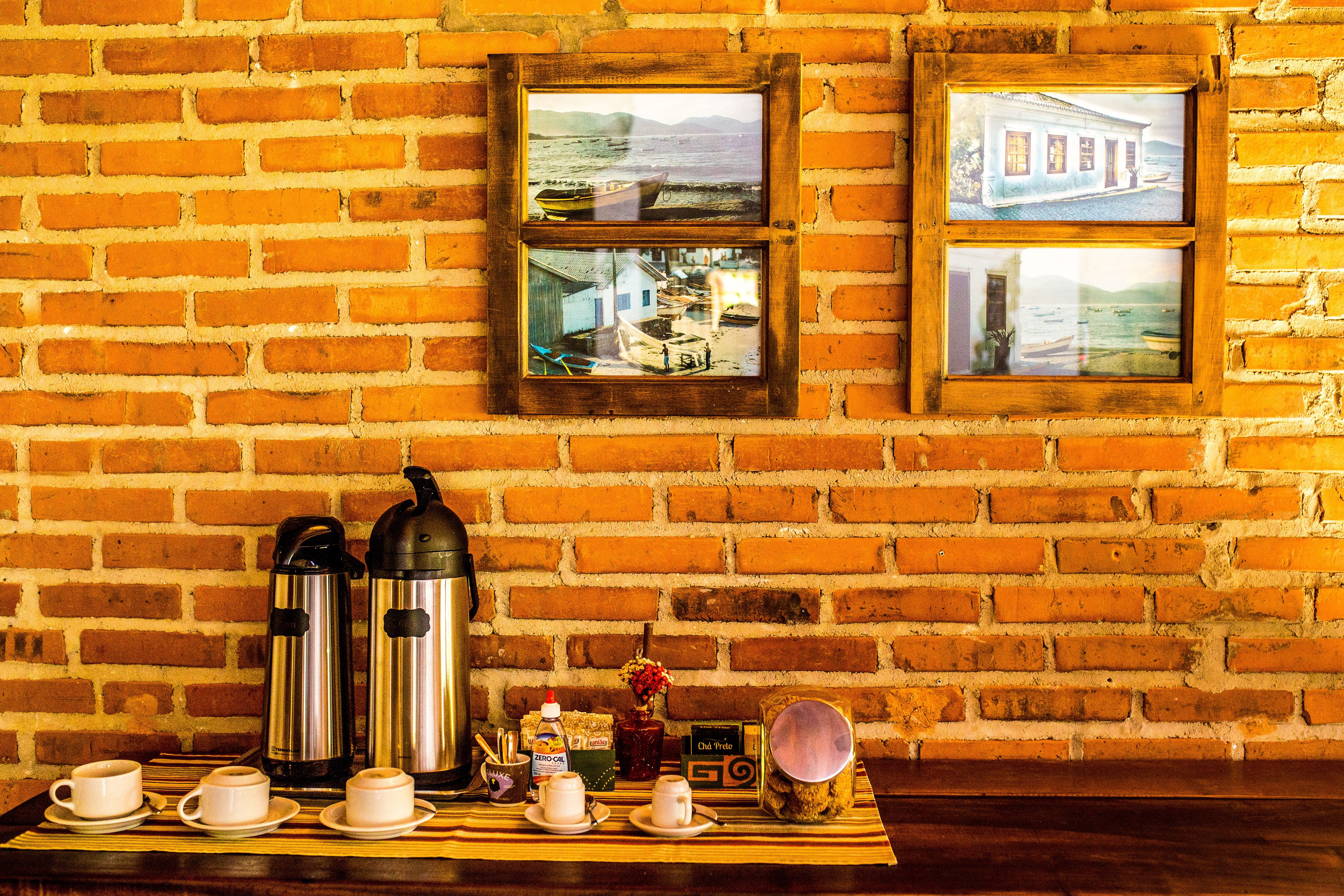 A brick wall with two framed pictures depicting coastal scenes, a table with coffee cups, thermos flasks, and a small clock.