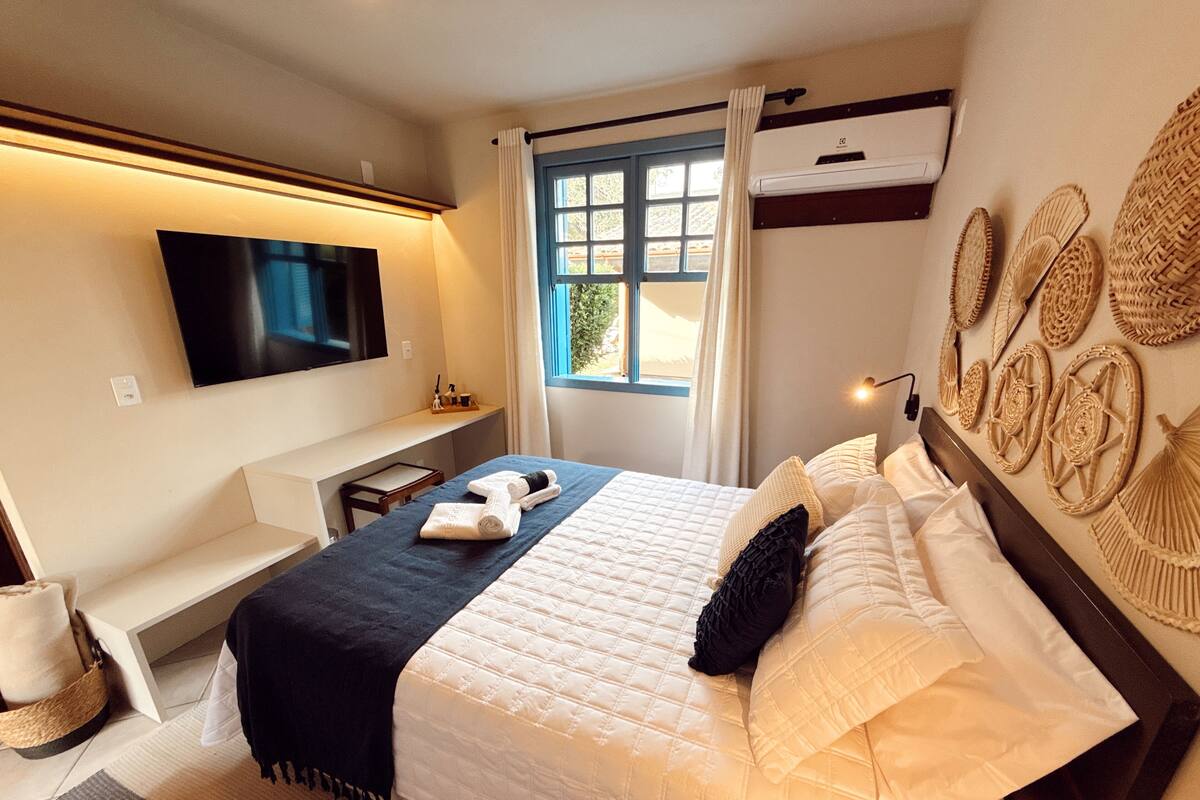 A hotel room with a bed, a TV mounted on the wall, a window with curtains, and a wall-mounted air conditioner.