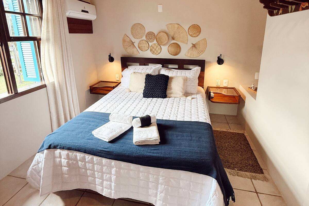 A bedroom with a wooden ceiling, a bed with a white quilt and blue throw, a wall-mounted air conditioner, and a bedside table with a lamp.