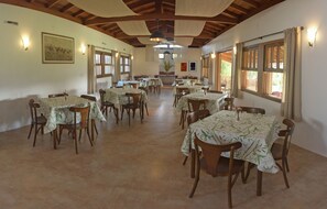 A dining area with multiple tables and chairs, wooden beams on the ceiling, and large windows with curtains. - Pousada da Lagoa (Garopaba)