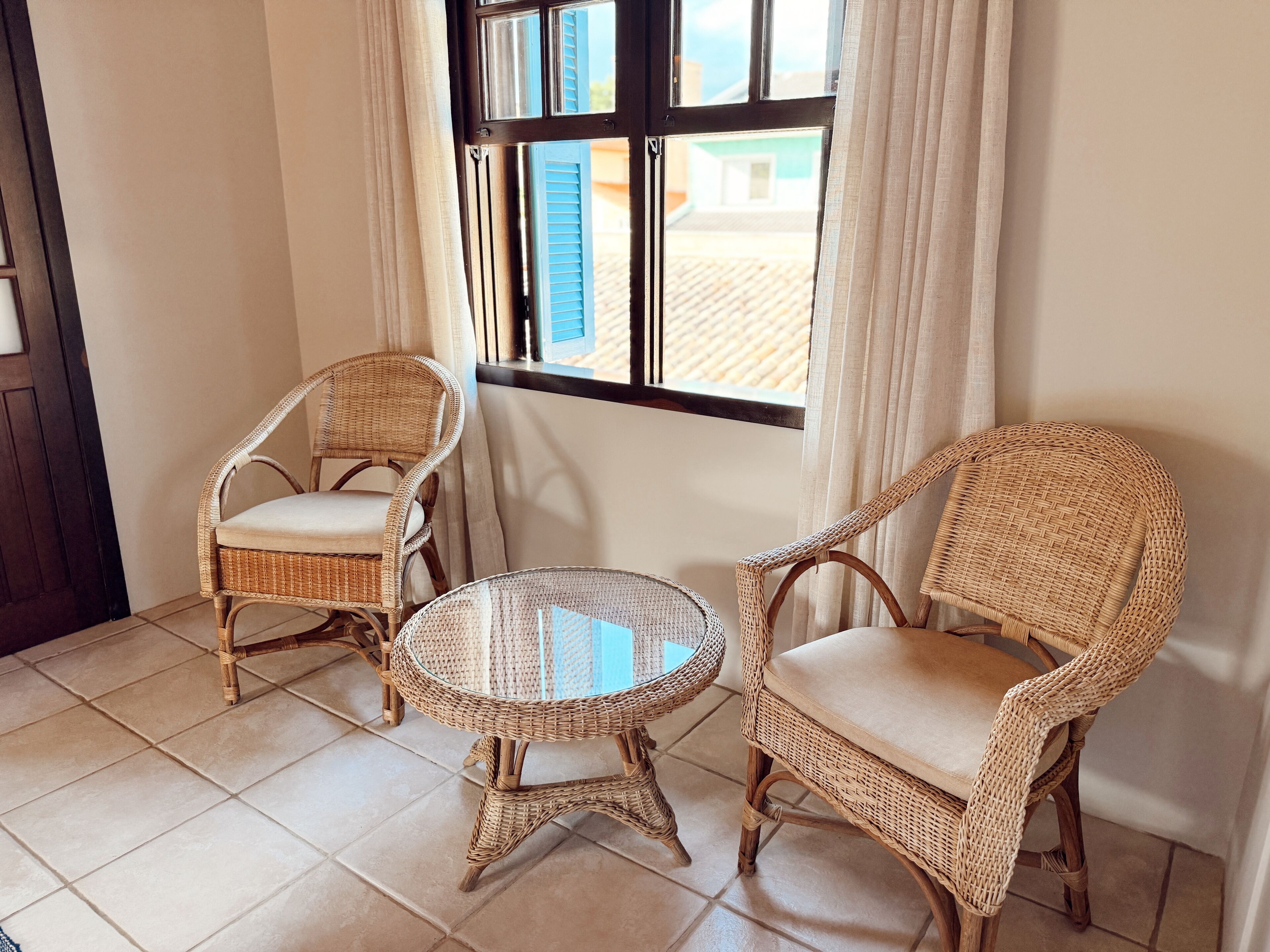 Una habitación con dos sillones de mimbre y una pequeña mesa redonda, una ventana con cortinas y suelo de baldosas.