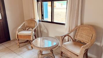 Una habitación con dos sillones de mimbre y una pequeña mesa redonda, una ventana con cortinas y suelo de baldosas.