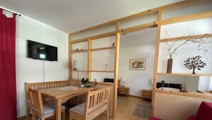 Ein Zimmer mit Esstisch aus Holz, Stühlen, einem an der Wand befestigten Fernseher, einem roten Sofa und einer Holzwand mit Regalen.