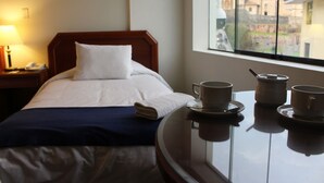 A hotel room with a bed, a bedside table with a lamp, and a window with a view of buildings. - Qarmenqa Hotel (Cusco)