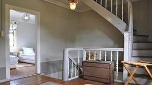 Un escalier avec des rampes blanches mène à une chambre à coucher où se trouvent un lit et une table de chevet. Un coffre en bois et une chaise pliante sont placés près de l’escalier.