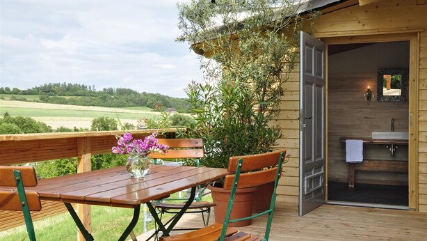 Une terrasse en bois avec une table et des chaises, un vase rempli de fleurs et une vue sur un paysage verdoyant.