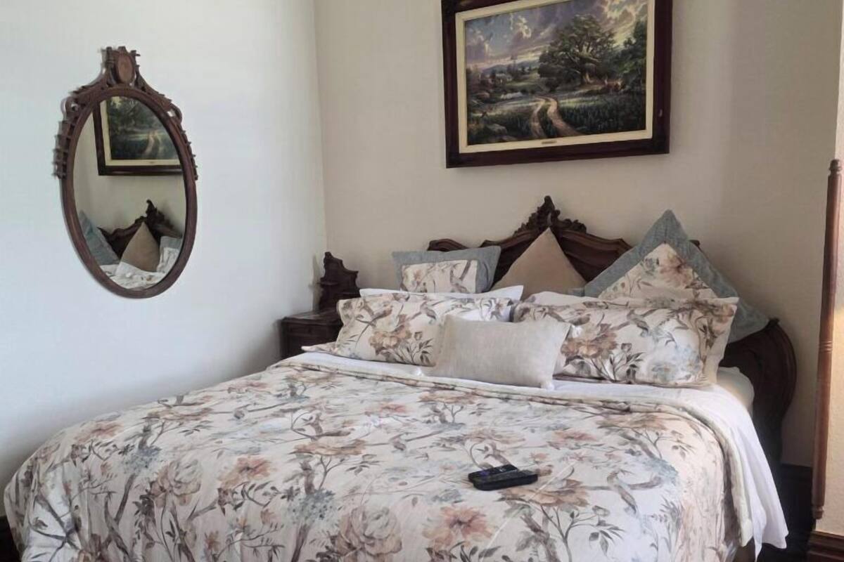 A bedroom with a floral bedspread, a wooden headboard, a mirror, and a landscape painting on the wall.