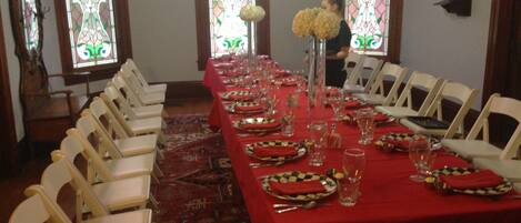 Un comedor con una larga mesa dispuesta para una comida, sillas blancas y vidrieras.
