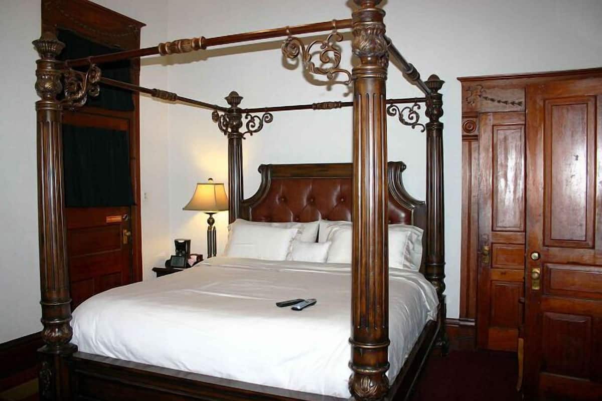 A four-poster bed with a wooden headboard and footboard, a bedside table with a lamp, and a wooden door in the background.