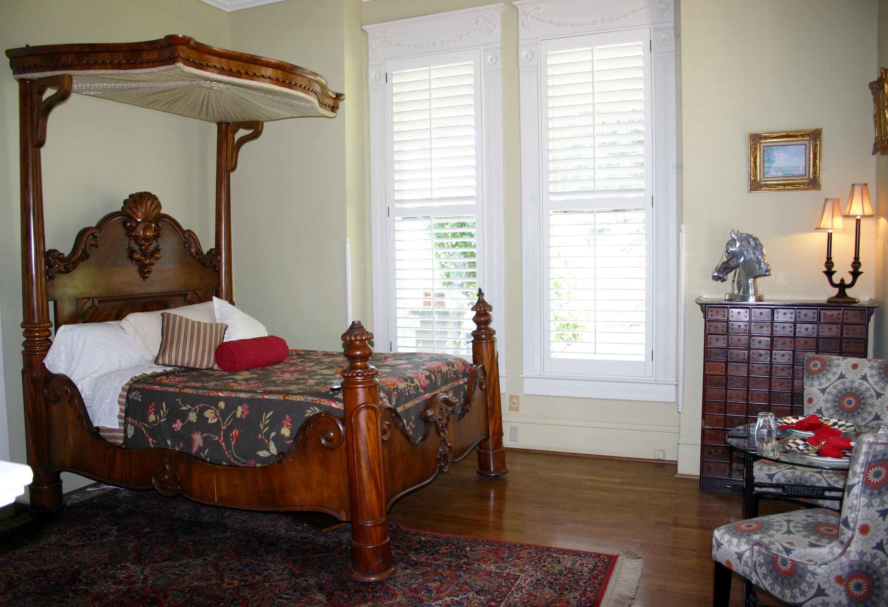 A four-poster bed with a canopy, a patterned bedspread, and a red pillow. There is a chair with a patterned cushion and a small table with a lamp.