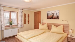A bedroom with a double bed, a bedside table with a lamp, a window with curtains, a radiator, and a wall-mounted picture of a deer. - Ferienwohnung 2 für 2 Personen mit Balkon, 49 m² (Inzell)