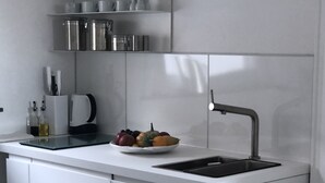 A modern kitchen with a white countertop, a sink, and a bowl of fruit. There are shelves above the countertop holding glasses and a kettle. - CITY FLAT Center Freudenstadt opposite Kurhaus  (Freudenstadt)