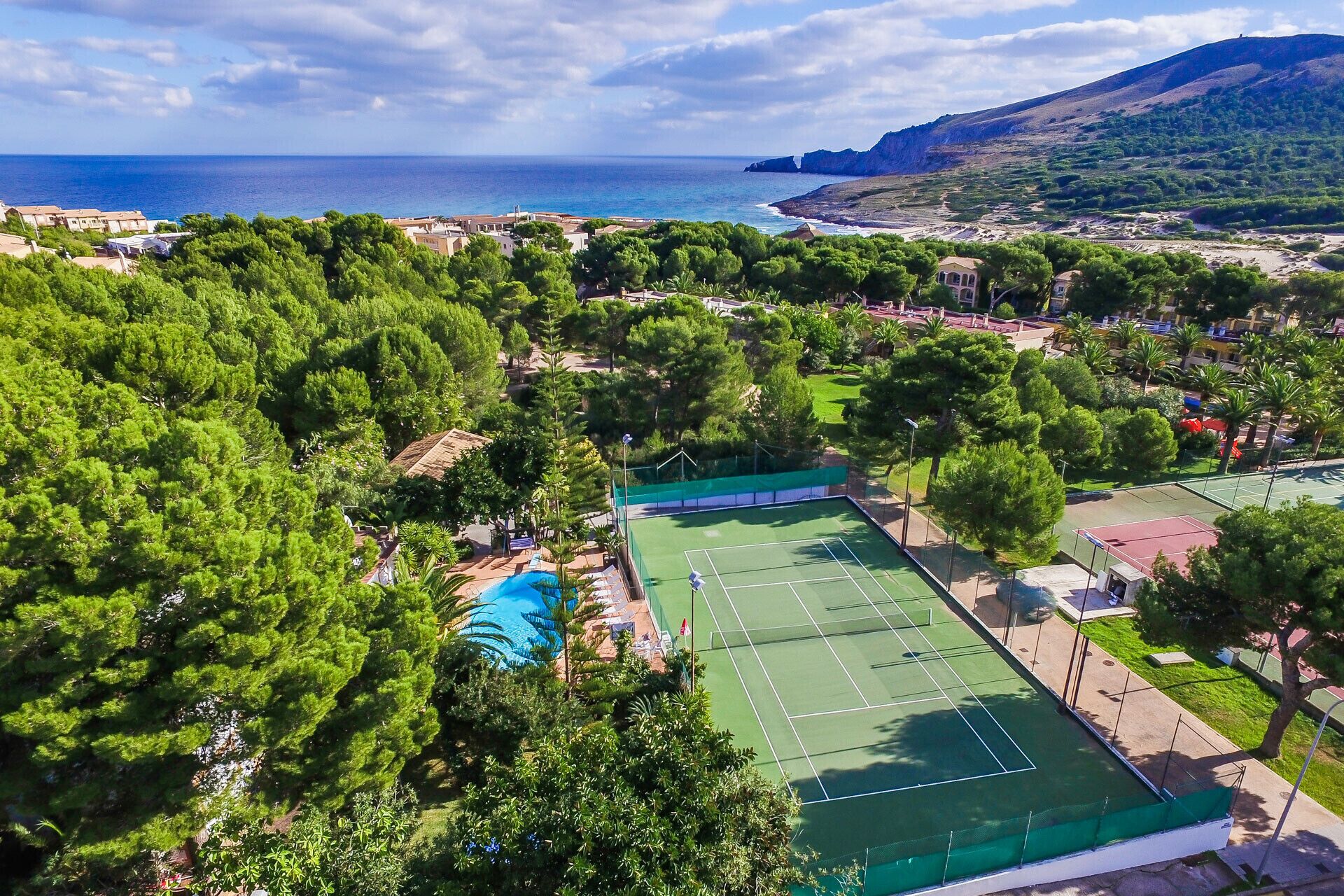Luchtbeeld van een kustgebied met een tennisbaan, een zwembad en weelderige begroeiing.