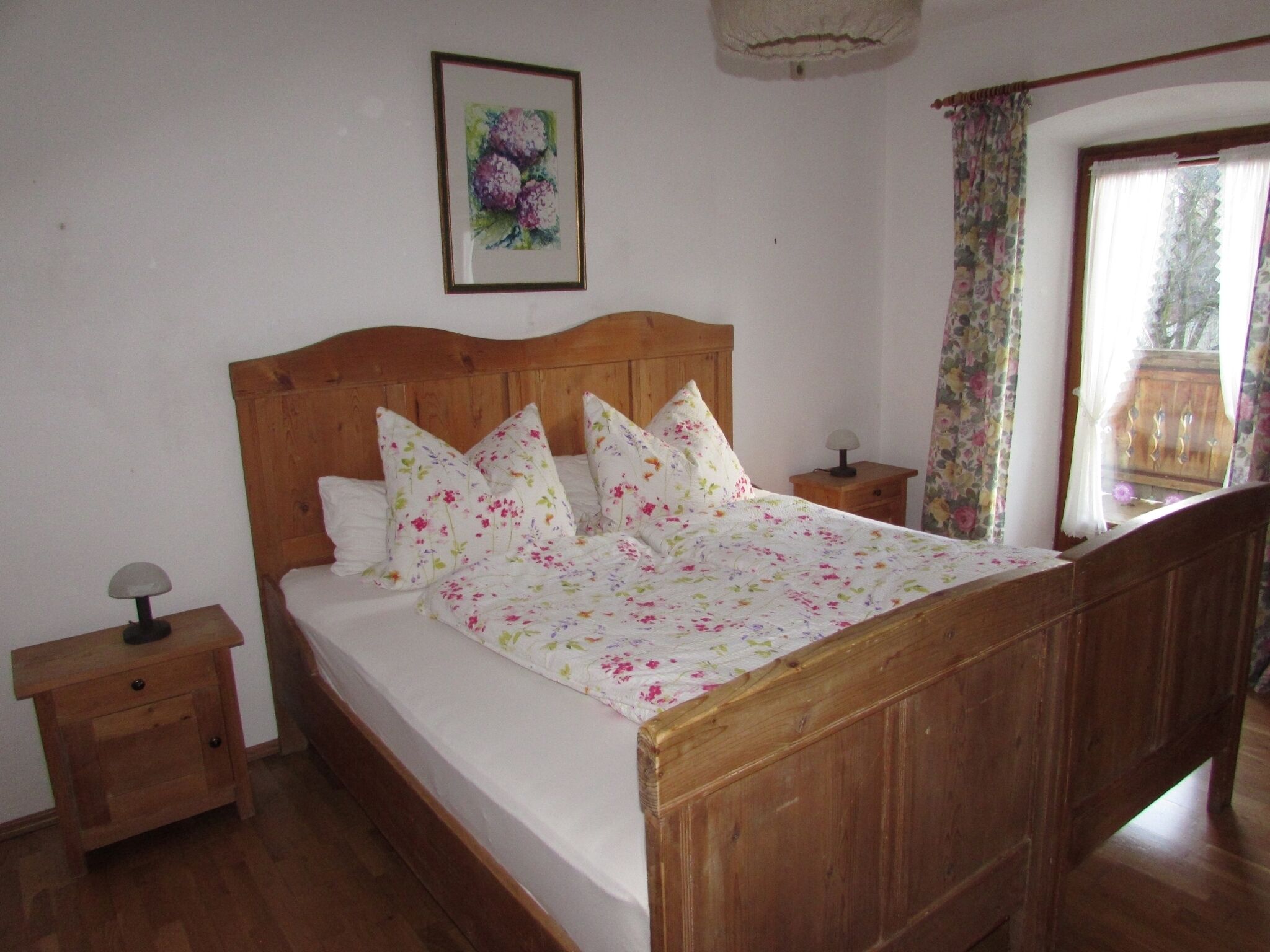 Un dormitorio con cama de madera, ropa de cama floral, una mesita de noche con lámpara y una ventana con cortinas.