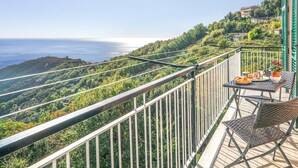 A balcony with a table set for two, overlooking a coastal landscape with a view of the sea and hills.