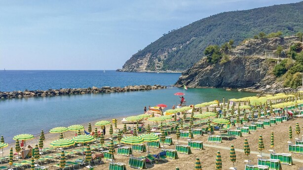 A beach with numerous green and yellow striped umbrellas and beach chairs, a rocky coastline, and a sailboat in the distance.