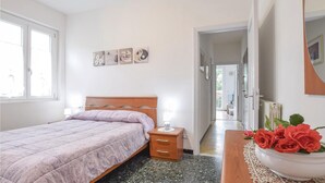 A bedroom with a bed, bedside table, and a nightstand with a lamp. There is a mirror on the wall and a door leading to another room.
