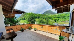 Ein Balkon mit Blick auf ein grünes Tal und Berge. Ein Holztisch und Kübelpflanzen sind vorhanden.