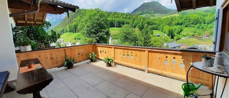 A balcony with a view of a green valley and mountains. There is a wooden table and potted plants.