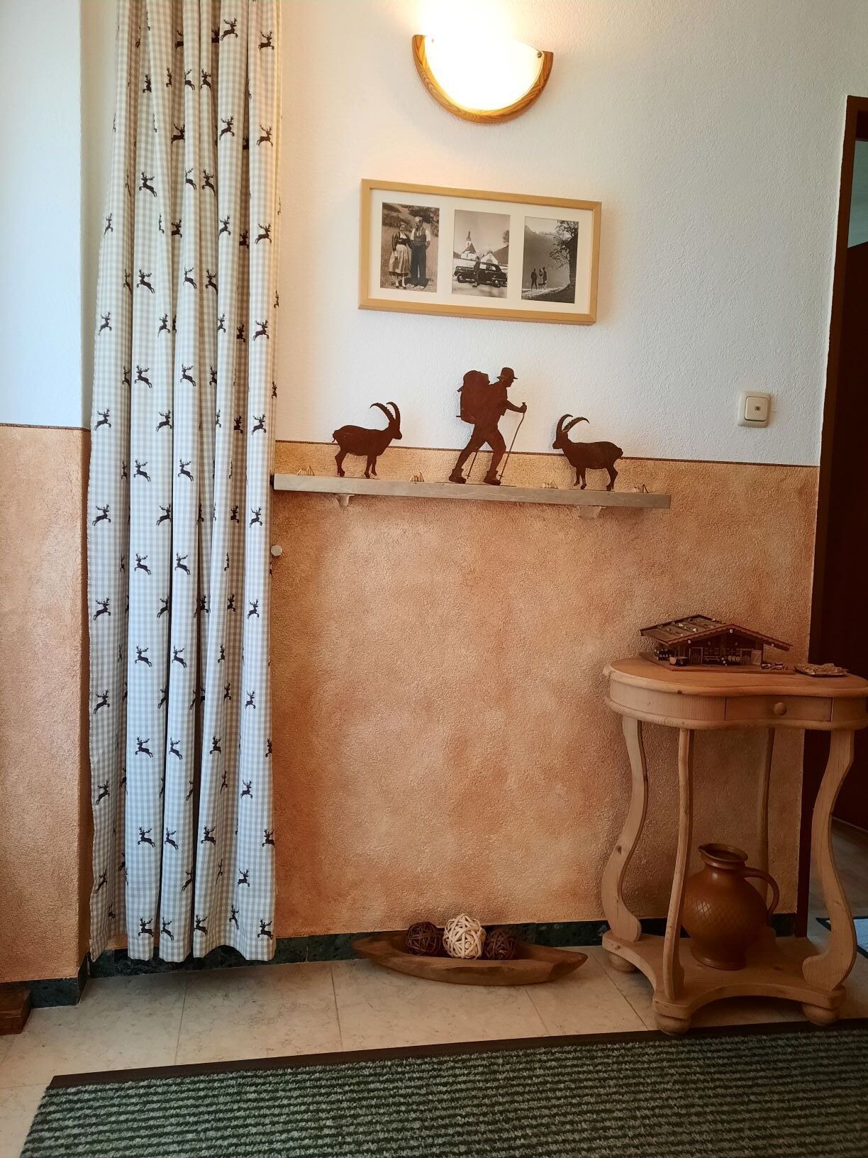 Huoneessa on ruutukuviollinen verho, seinään kiinnitetty valaisin, pienoismallien koristama hylly, pieni pöytä, jossa on maljakko, ja matto.