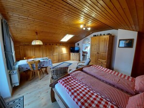 Ein gemütliches Zimmer mit Holzbalkendecke, einem Bett mit karierter Bettwäsche, einem Esstisch mit Stühlen und einem Fernseher.