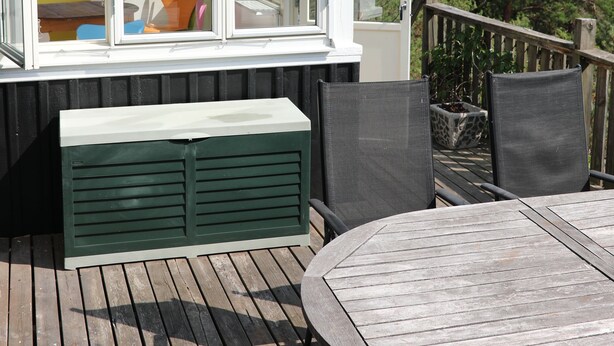 Una terraza de madera con una mesa redonda y sillas, una ventana con vista a muebles de exterior, y una barandilla de madera.