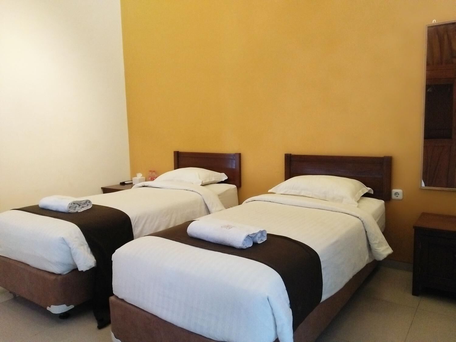A hotel room with two single beds, each with a wooden headboard, white bedding, and a rolled towel on the bed.