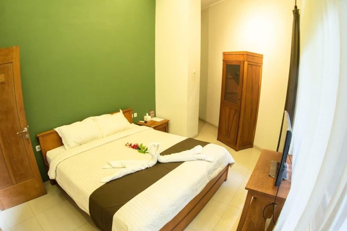 A neatly made bed with white linens and a brown bedspread, a wooden nightstand with a lamp, a wooden wardrobe, and a television on a wooden table.