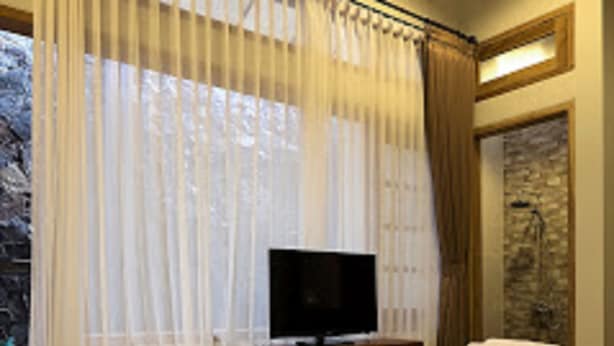 A bedroom with a bed, a television on a wooden nightstand, and a stone wall behind the bed. - Helena Guest House (Malang)