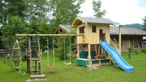A wooden playhouse with a slide and swings in a grassy yard. - Vacation apartment 2 Kienbergblick, 4 persons, 1 separate bedroom, bed-sitting room, 36 sqm (Inzell)