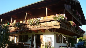 Un chalet traditionnel en bois, doté d’un balcon orné de fleurs, d’un porche en bois et surplombant un ciel d’un bleu éclatant.