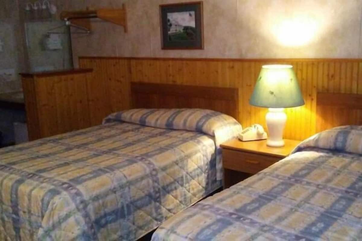 A room with two beds, wooden headboards, a nightstand with a lamp, and a wall-mounted shelf with a picture.