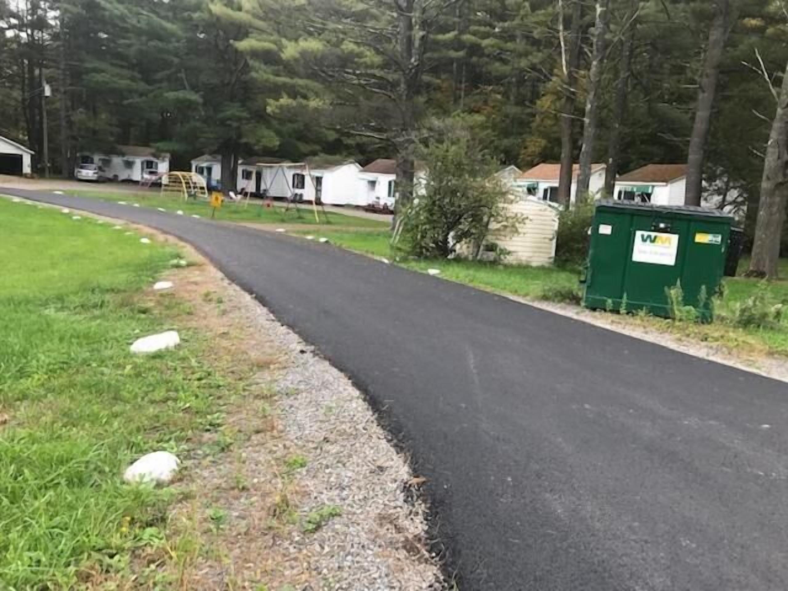 A paved road with a grassy median, surrounded by trees and a row of mobile homes.