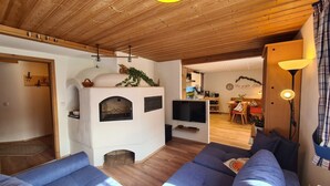 A cozy living room with a fireplace, a television, and a dining area in the background. - Ferienwohnung Nr. 1, 1-2 Personen, 60 qm, Dusche/WC, Terrasse, TV, W-LAN, 1 Schlafzimmer + 1 Wohnschlafzimmer, Kachelofen (Ramsau bei Berchtesgaden)